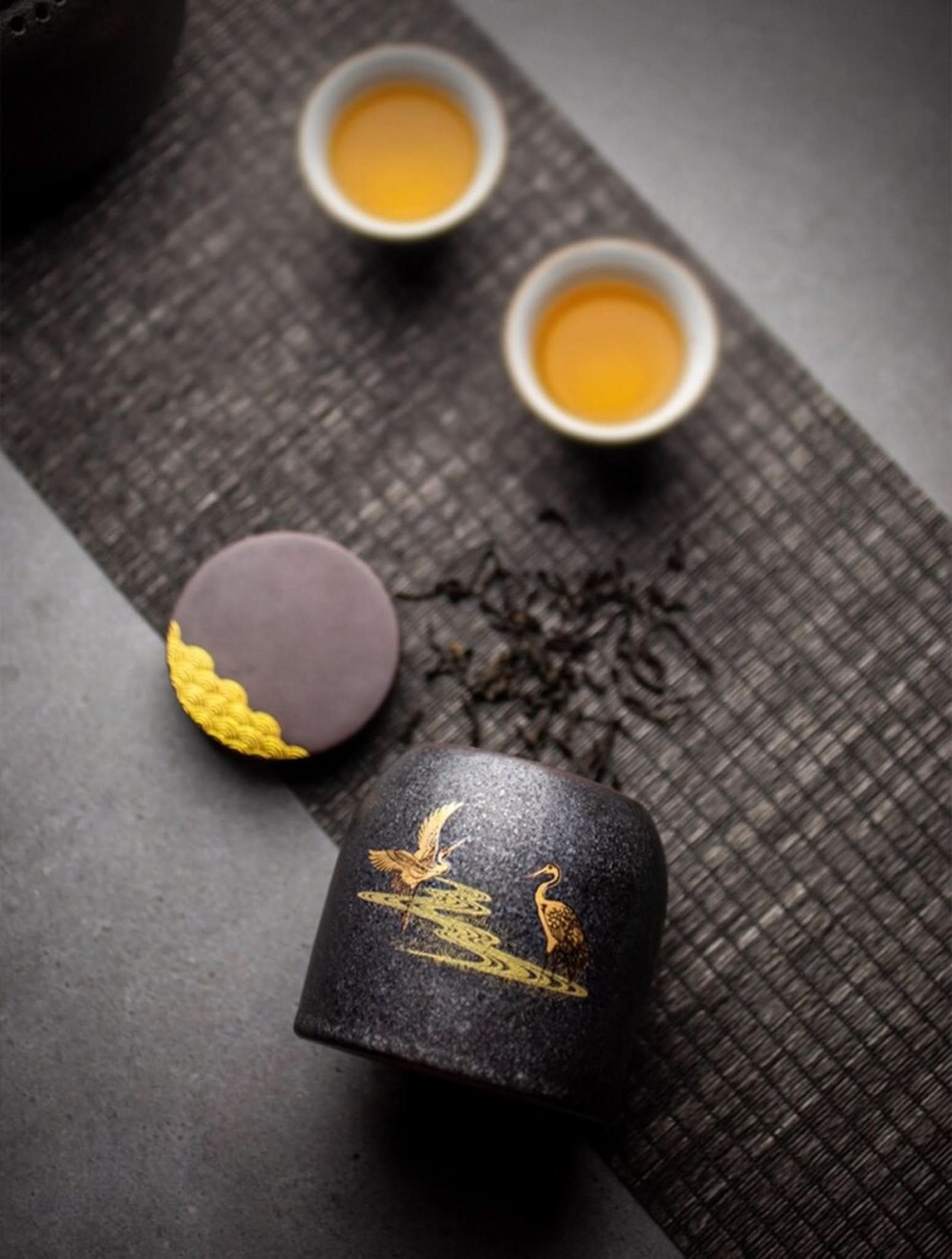 Retro black ceramic tea canister with golden cranes, on a textured mat with tea and cups.